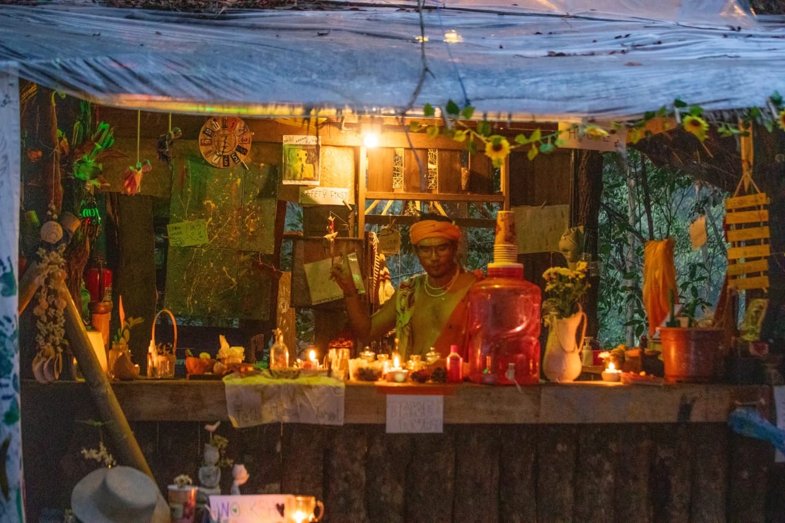 A camp bar built from scrap timber and tarpaulin at dusk, hung with sunflowers, candles and hand-lettered signs.